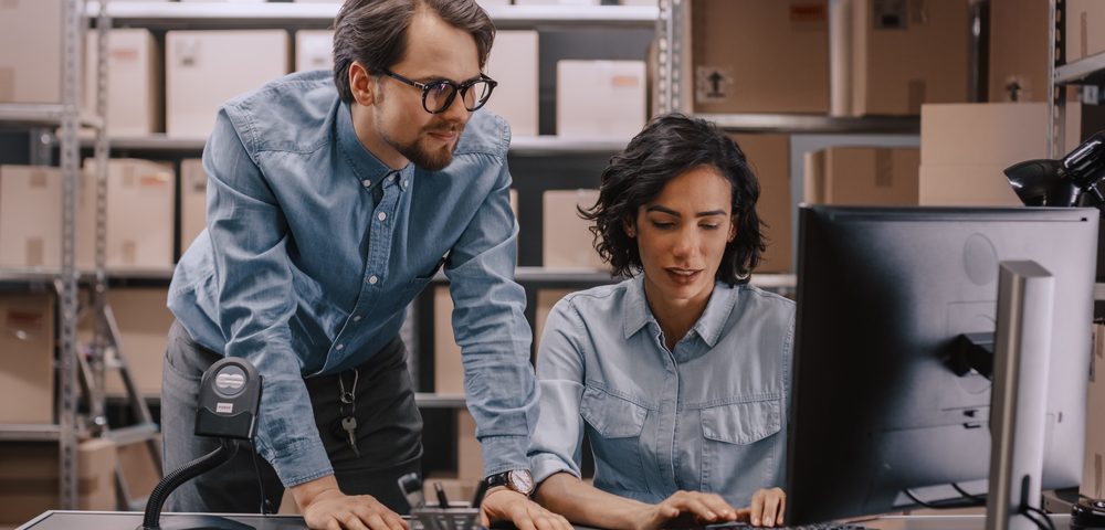 Male and Female Warehouse Inventory Managers Talking, Solving Problems, Using Personal Computer and Checking Stock. In the Background Rows of Shelves Full of Cardboard Box Packages.